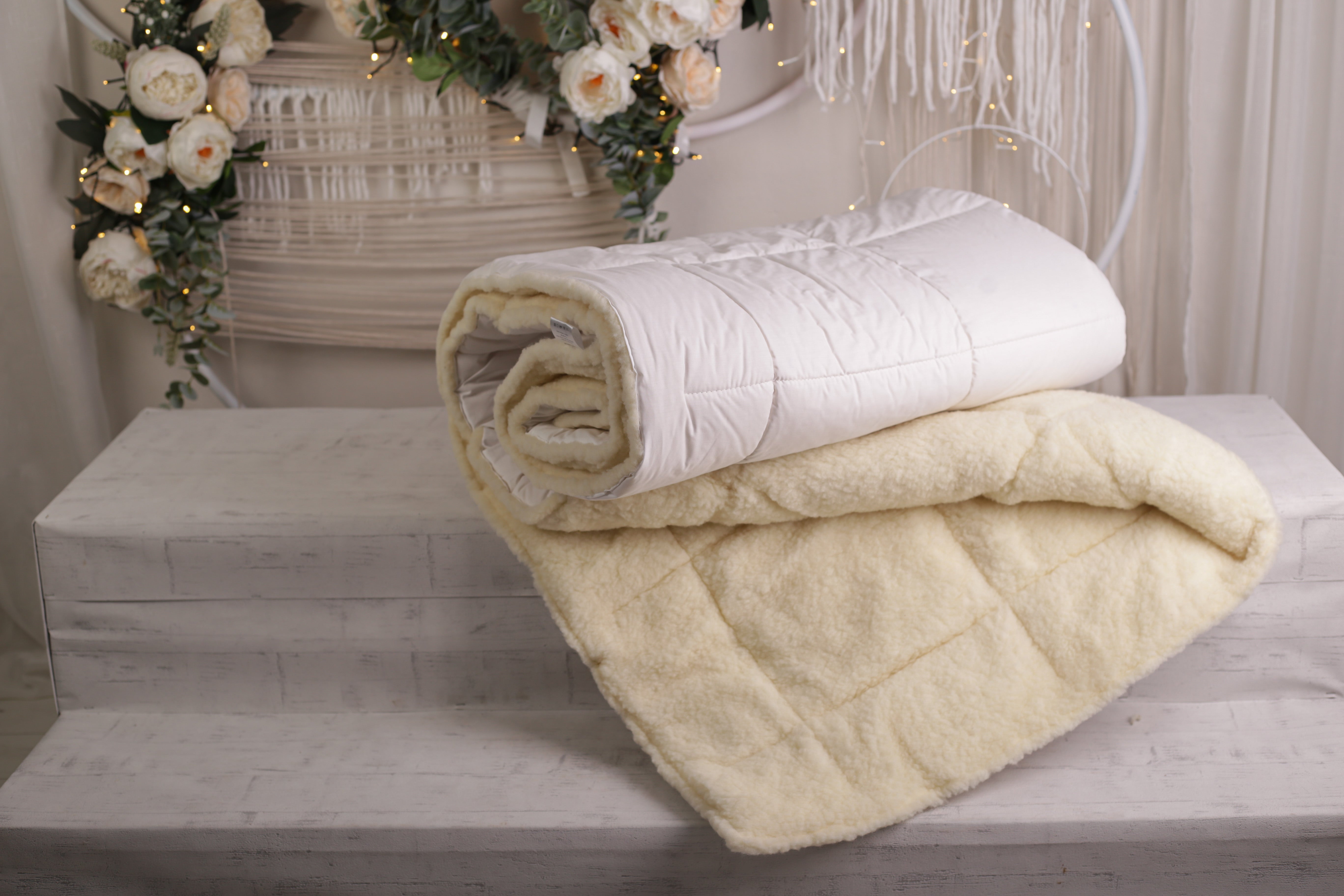 Die Bettdecke Gold Lux von Wool Line liegt eingerollt auf einer schönen Ablage mit Blumen im Hintergrund. Zum Vorschein kommt die eine Hälfe des Bezuges aus hochwertigem Wollfilt, während die andere Seite aus Bio-Baumwolle für kühlende Entlastung im Sommer sorgt.