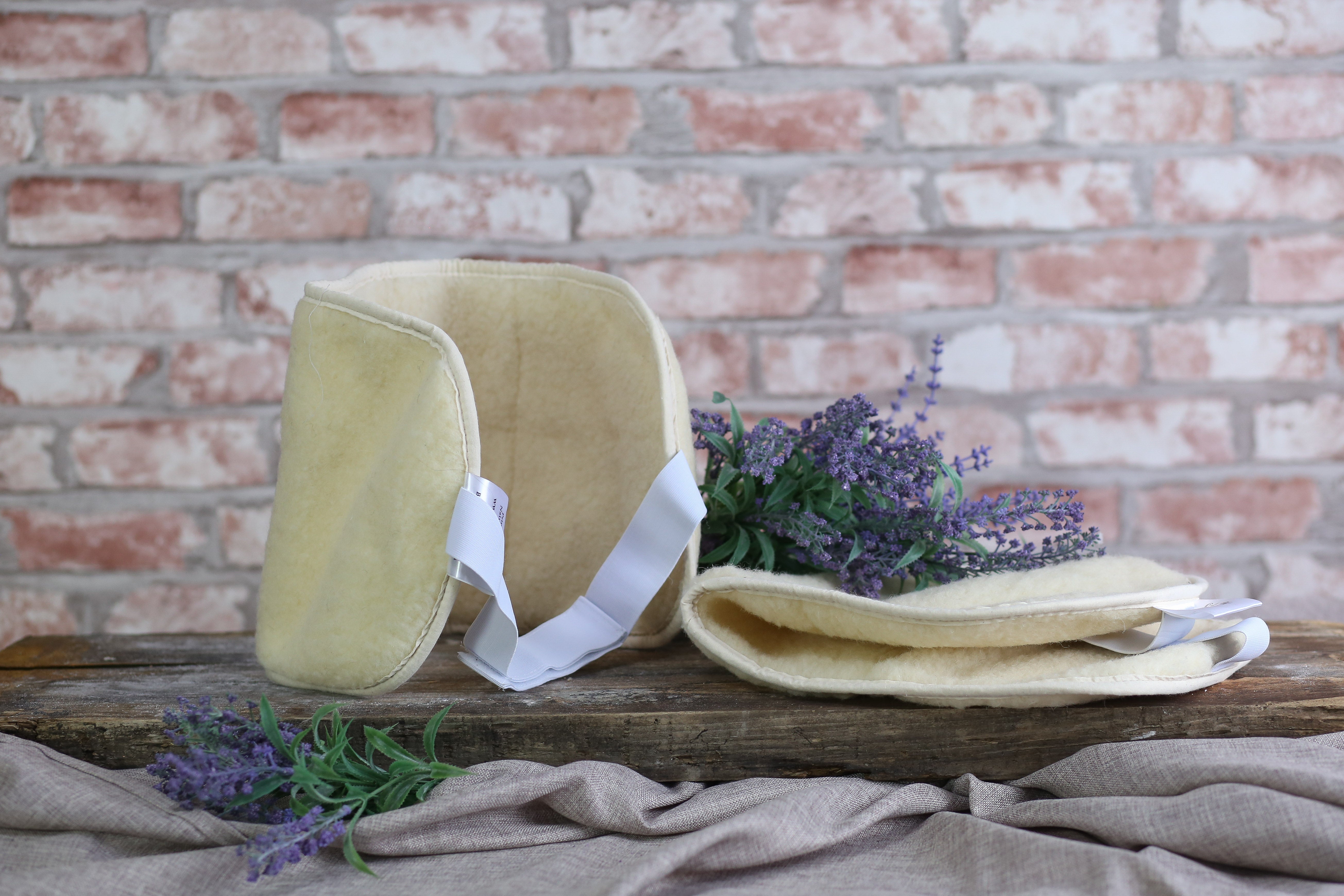 Zwei Bio-Rückengurte aus Naturwolle von Wool-Line auf einer kleinen Anhöhe aus dunkelbraunem Holz. Der linke Rückengurt ist aufgestellt und ausgeklappt, während der rechte Rückengurt eingeklappt danebenliegt. Dazwischen sind zwei lila Pflanzen zur Dekoration aufgestellt.