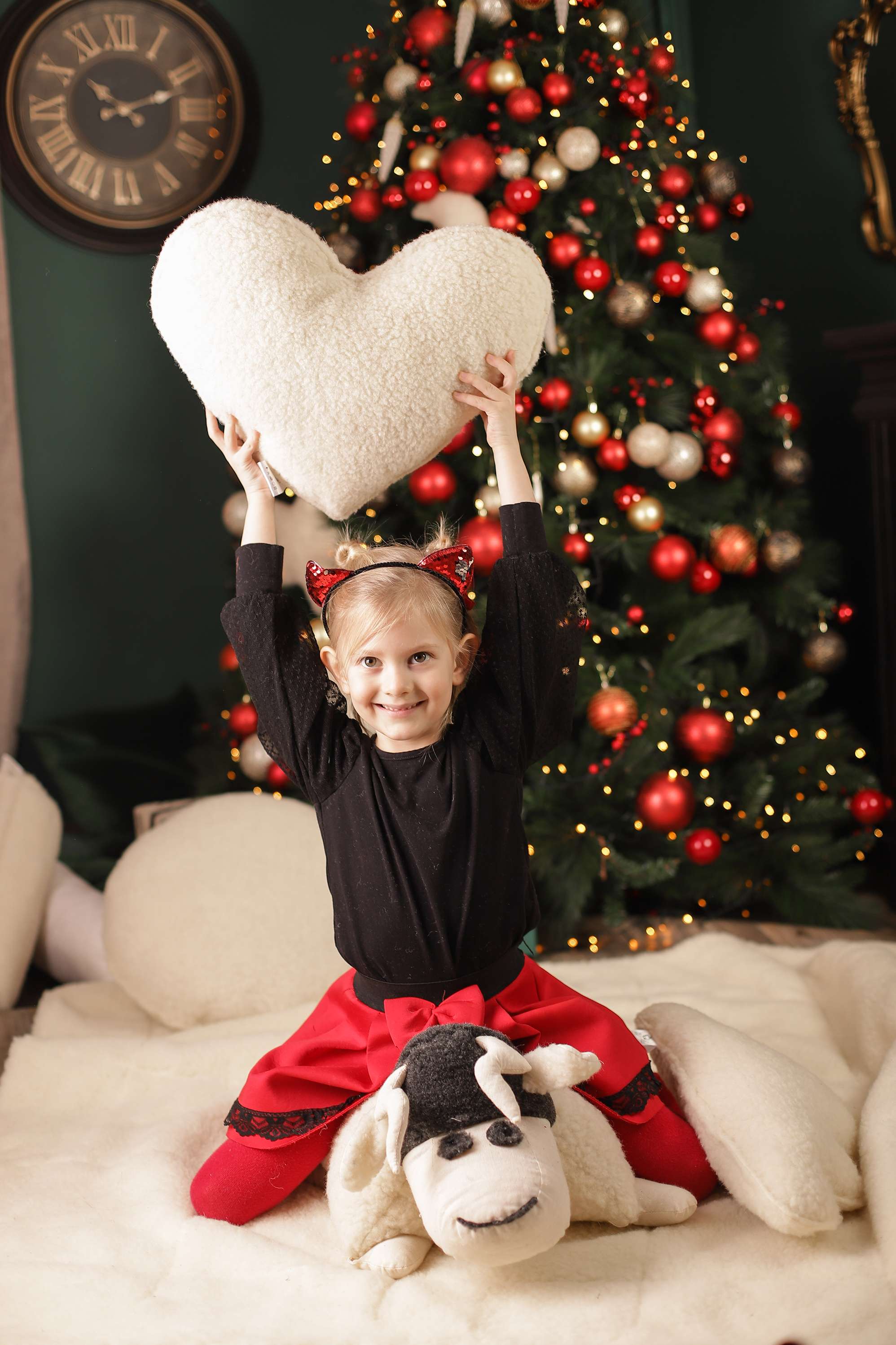 Ein Mädchen sitzt vor einem bunt geschmückten Weihnachtsbaum und hält glücklich das Herz-Kuschelkissen von Wool-Line in die Höhe. Es scheint so, als habe sie das Herzkissen zu Weihnachten geschenkt bekommen.
