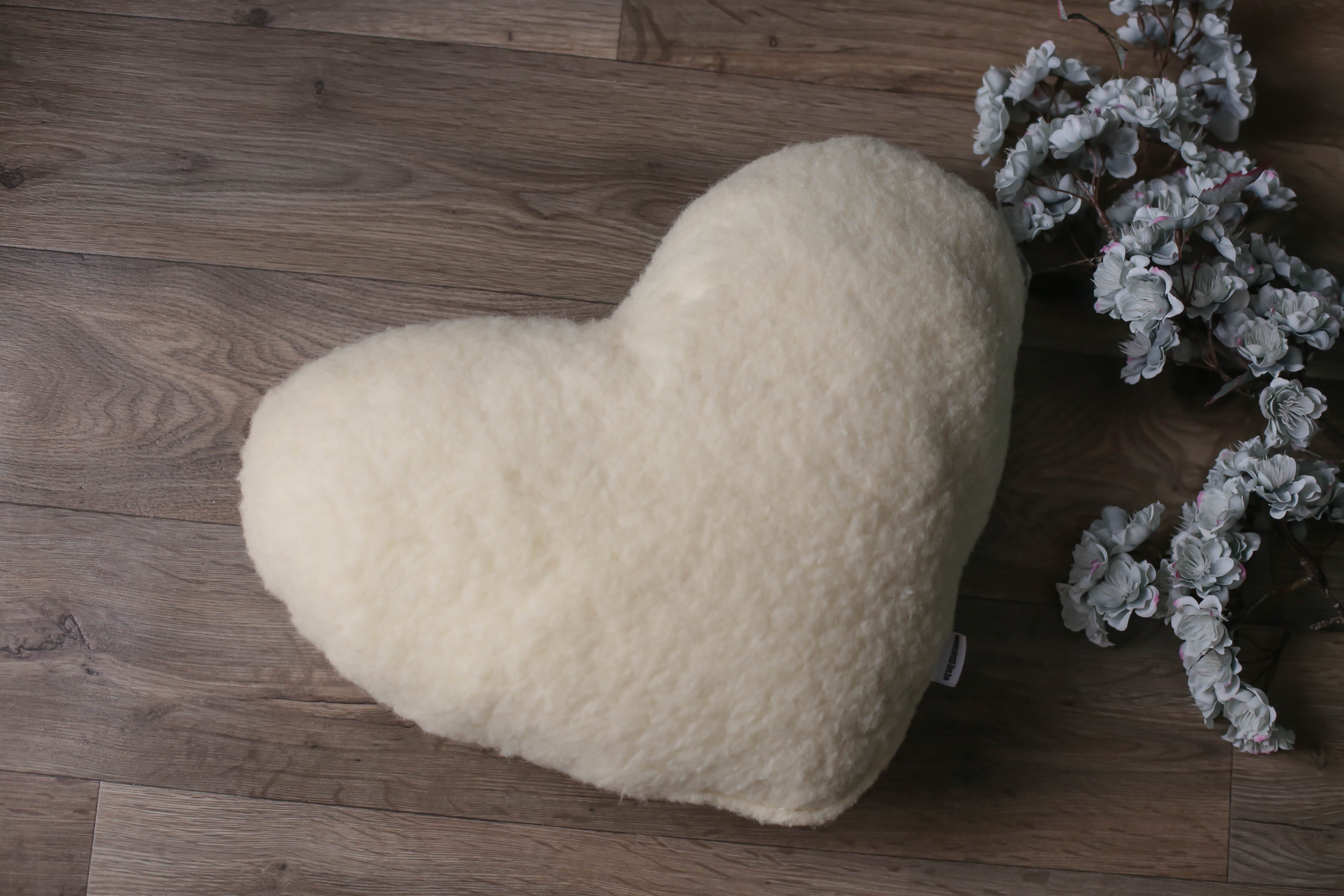 Ein herzförmiges Kissen von Wool-Line auf einem dunkelbraunen Holzfußoden abgelegt, Rechts daneben liegen dezente weiße Blüten zur Dekoration.