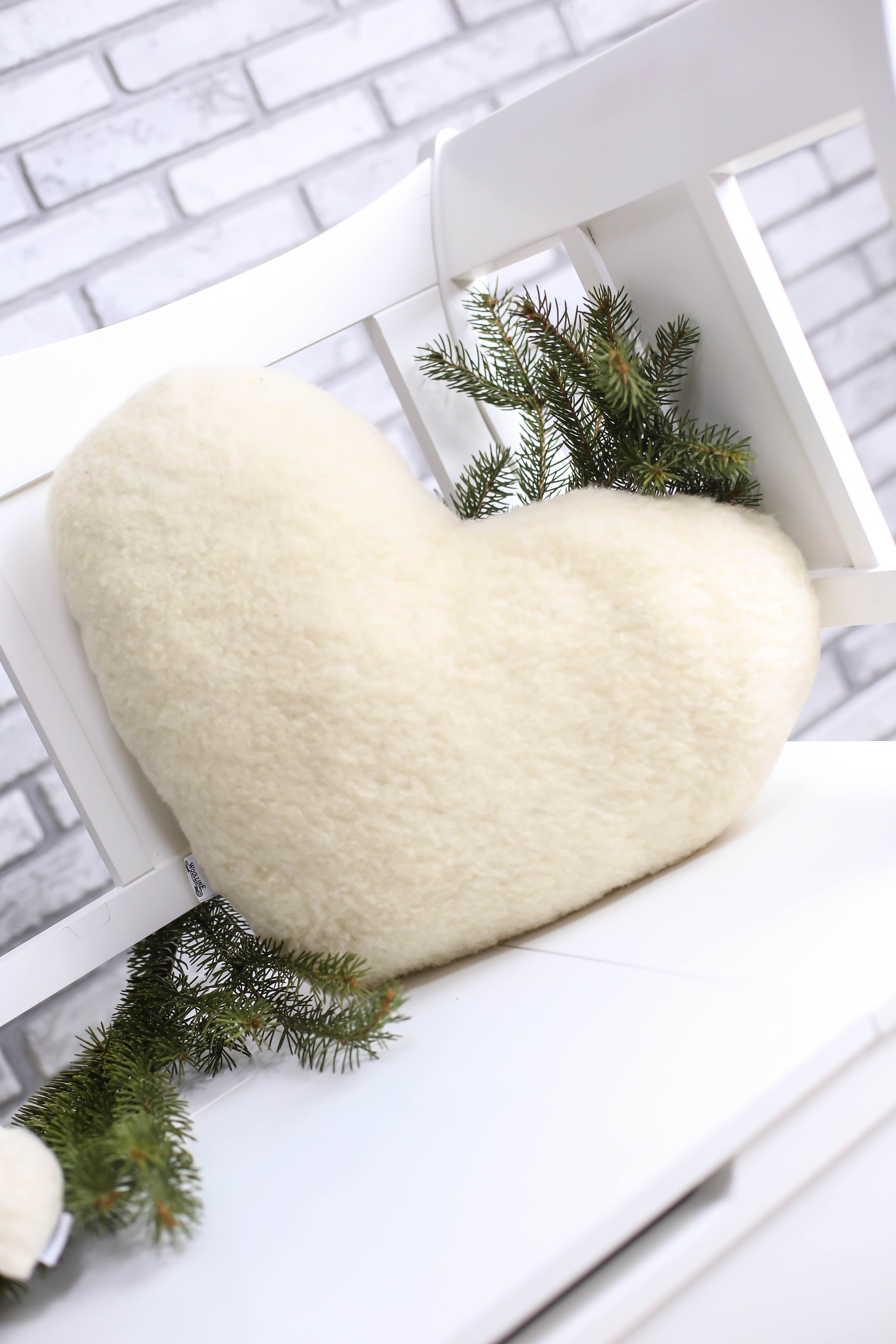 Das beige Herzform-Kissen aus Schafschurwolle dekorativ auf einer weißen Outdoor-Bank abgelegt. Grüne Nadelbaum-Äste sind zur weiteren Dekoration beigelegt und unterstreichen die Natürlichkeit des Herzkissens.