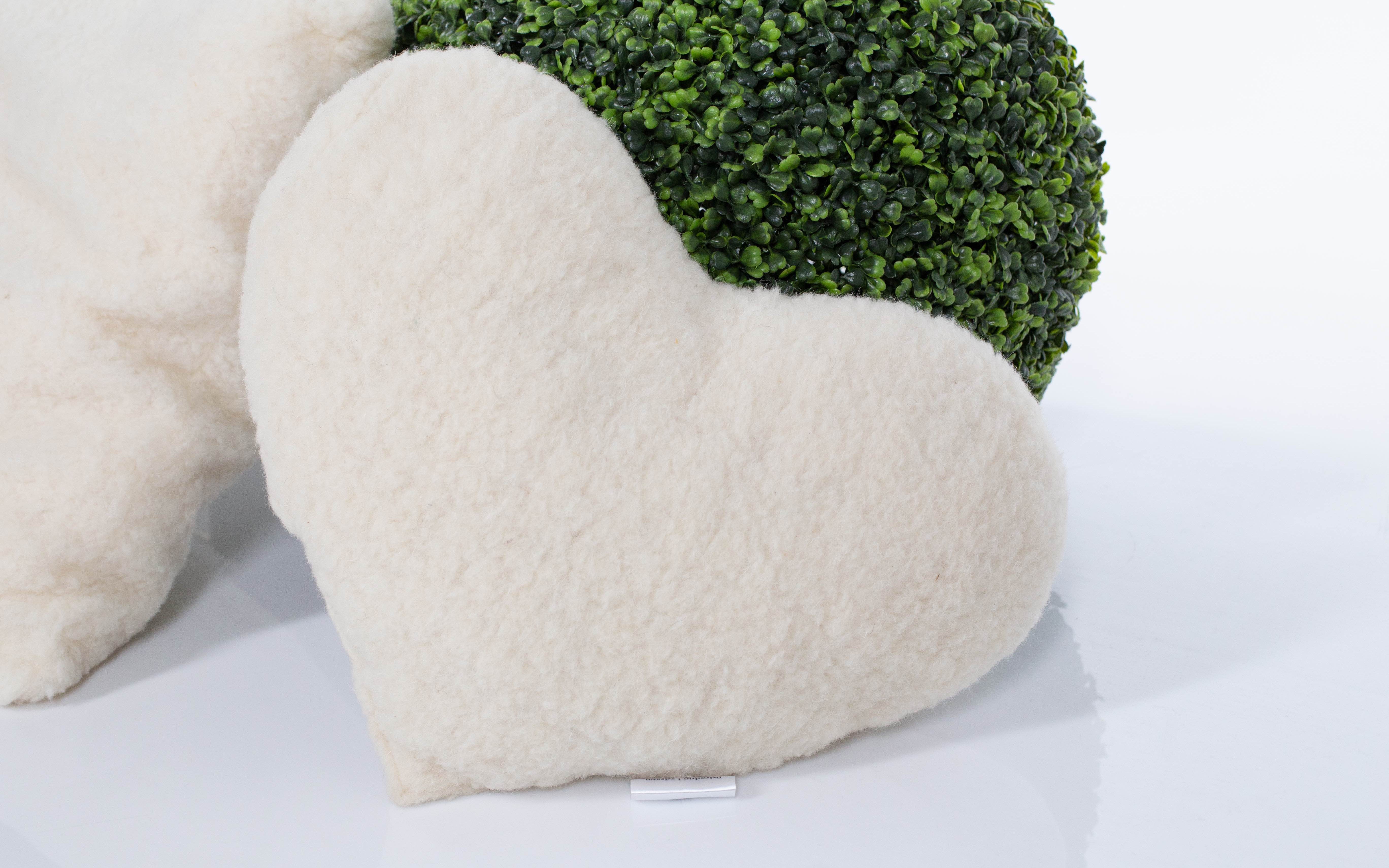 Das weiße Kuschelherz Kissen von Wool-Line auf weißem Untergrund fotografiert. Im Hintergrund befindet sich eine runde, güne Pflanze. Der weiche Filz des Herzkissens ist detailliert und gut ausgeleuchtet zu sehen.
