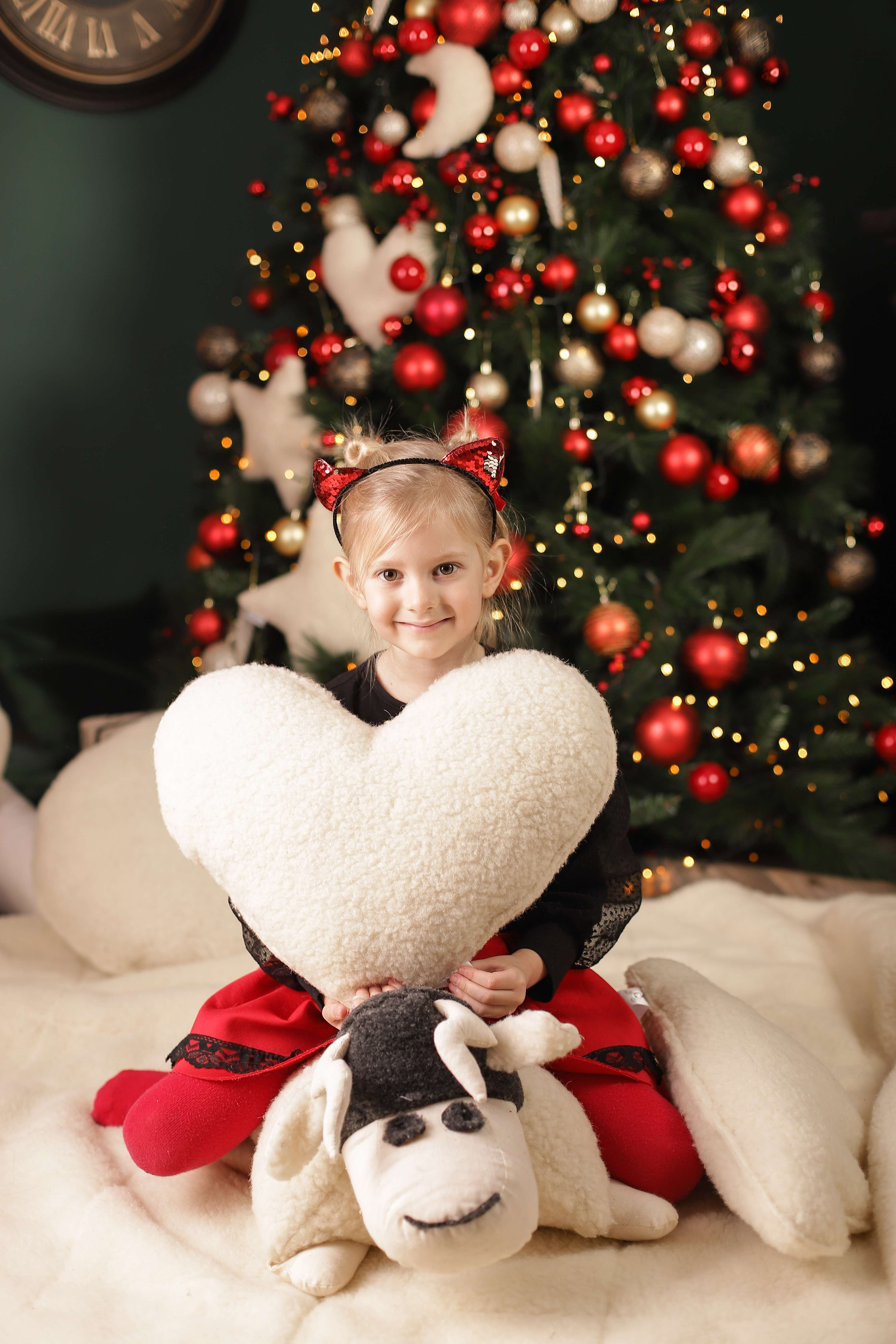 Das Kuschelkissen Herz von Wool-Line in den Händen eines glücklich lächelnden Mädchen, das zirka 10 Jahre alt ist. Im Hintergrund steht ein Weihnachtsbaum geschmückt mit roten, weißen und goldenen Kugeln. Das Mädchen hat das Herzkissen offenbar zu Weihnachten geschenkt bekommen.