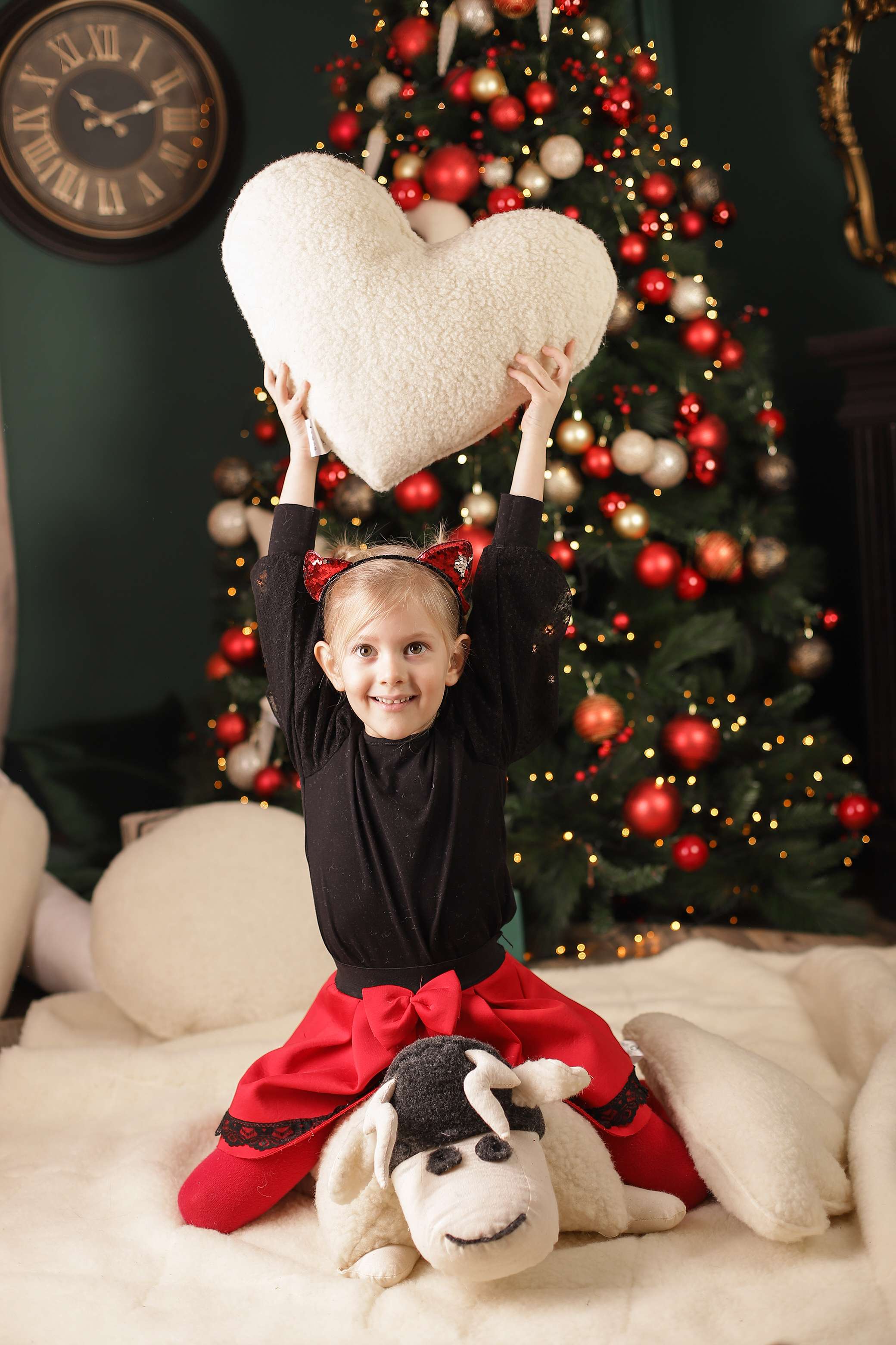 Ein etwa zehnjähriges Mädchen reckt das Kuschelkissen Herz von Wool-Line freudestrahlend vor einem bunt geschmückten Weihnachtsbaum in die Höhe.
