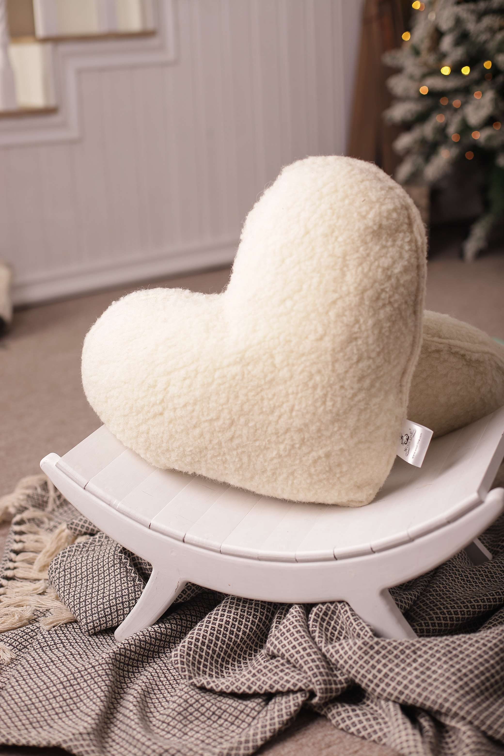 Das Kuschelkissen in Herzform von Wool-Line auf einem kleinen weißen Hocker aufgestellt. Unter der Anhöhe befindet sich eine kleine schwarz-weiß-karierte Decke.