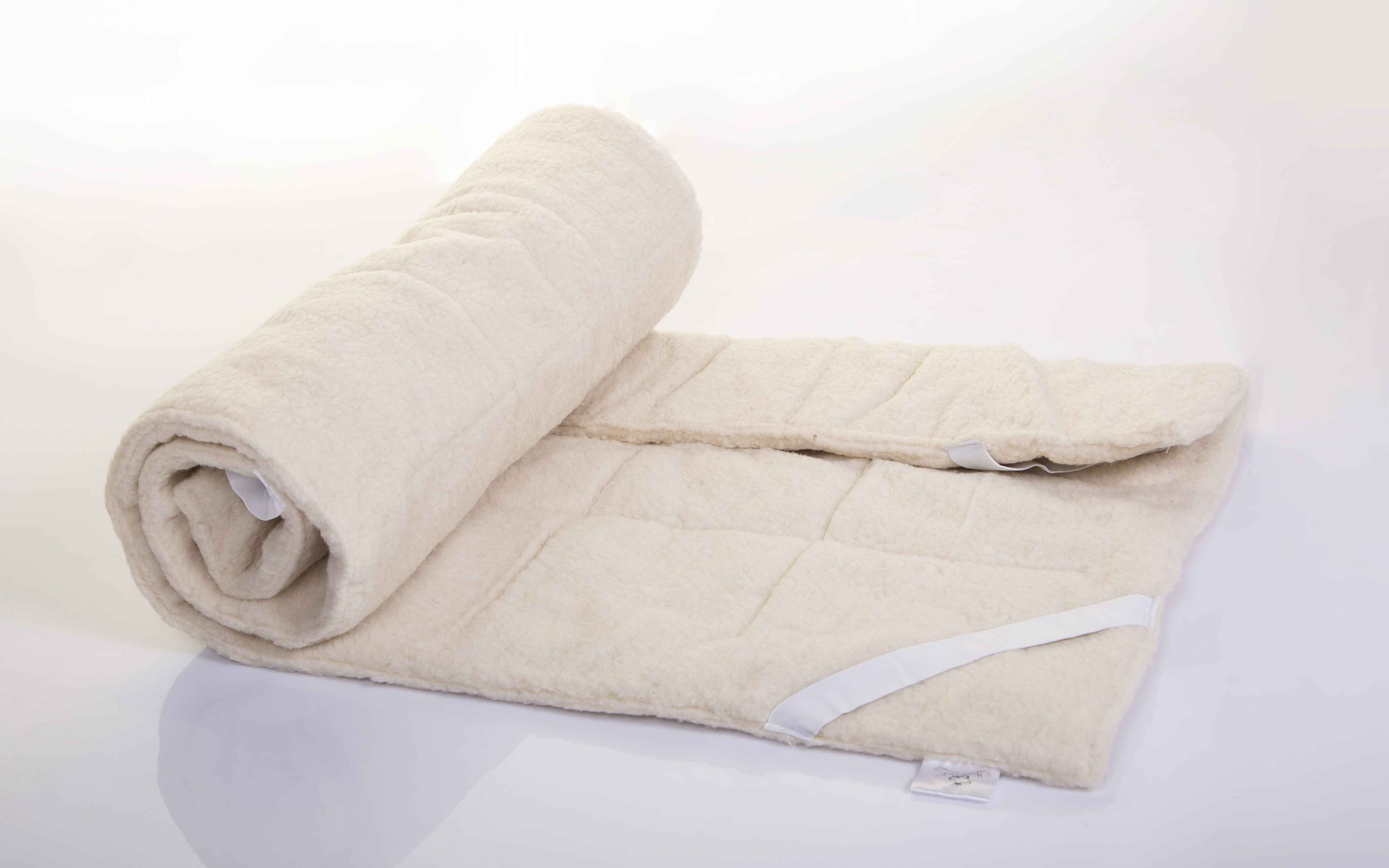 Die Matratzenauflage Schurwolle Gold von Wool-Line auf weißem Untergrund. Die linke Seite ist flexibel eingerollt, die andere Seite liegt teilweise zusammengelegt flach auf dem Untergrund. Dank der Schafschurwolle im Futter der Matratzenauflage ist das Produkt sehr elastisch ohne sichtbare Dellenbildung.