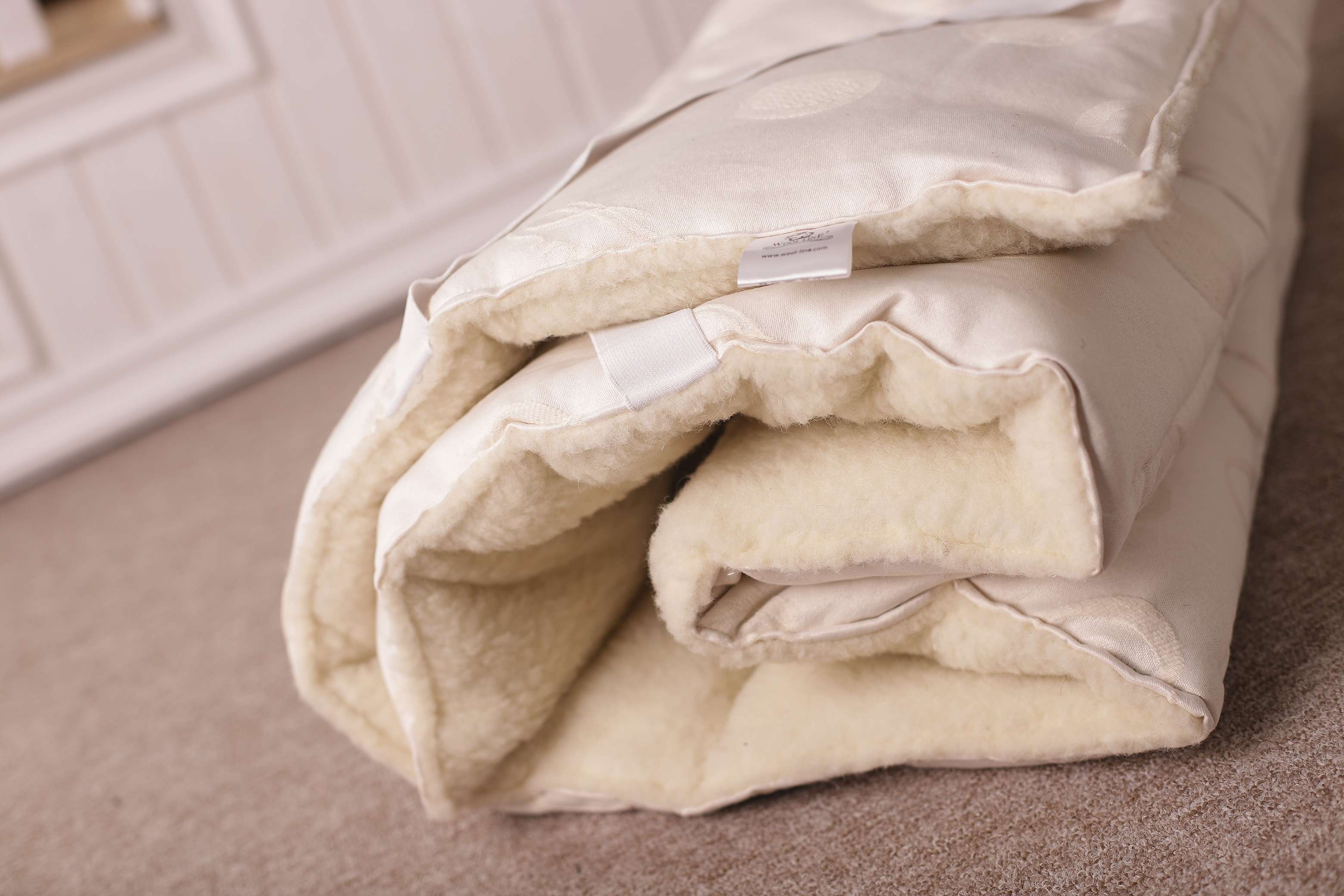 Der Matratzentopper Natur old/Lux von Wool-Line zusammengelegt auf dem Boden. An der Unterseite kommt der weiche Filzbezug aus Schafschurwolle genau zum Vorschein, der das ganze Jahr über die Temperatur auf dem Topper angenehm und natürlich reguliert.