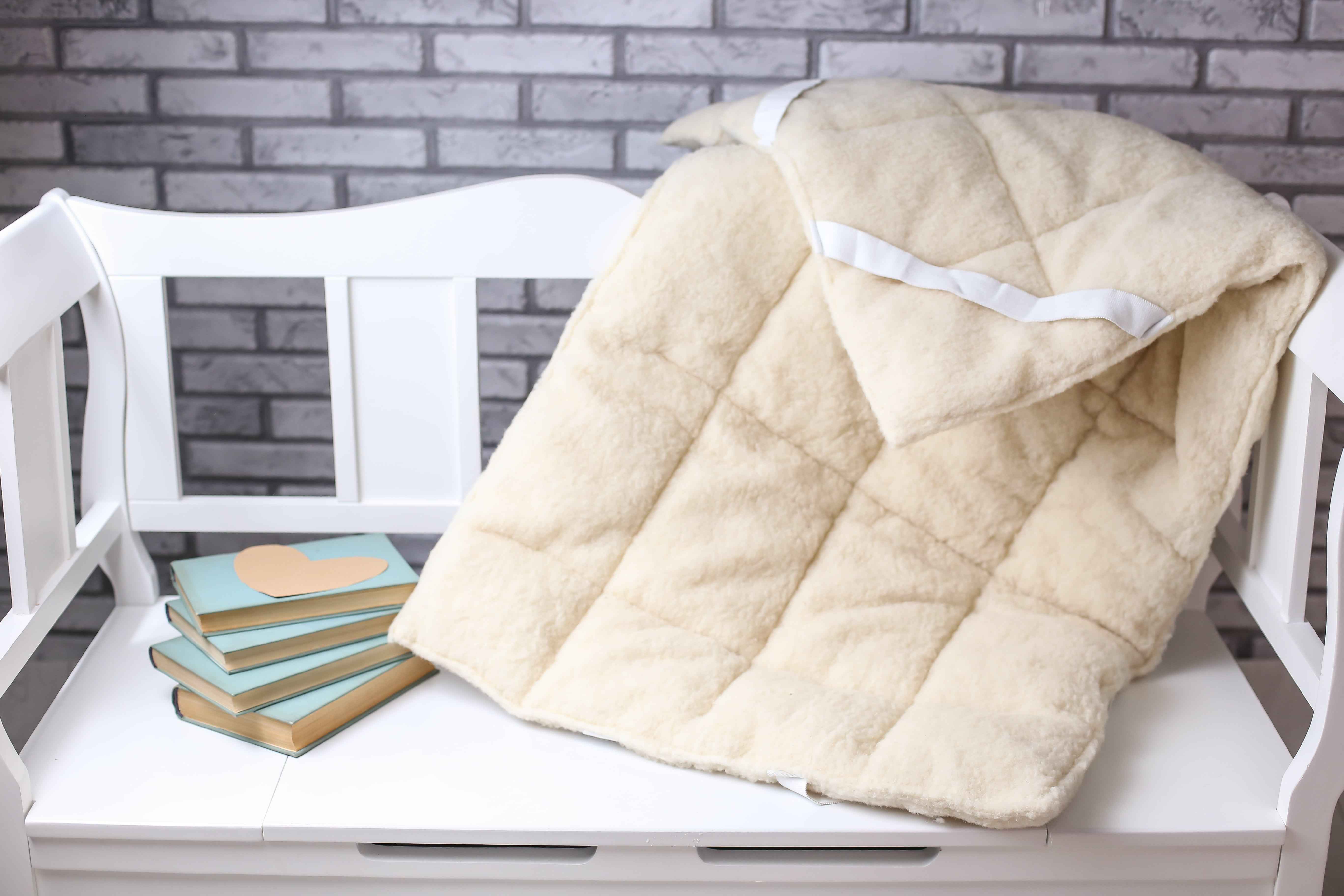 Die Schurwolle Matratzenauflage Gold von Wool-Line liegt auf einer weißen Bank. An der oberen Spitze ist einer der weißen Spanngurte zu sehen, mit denen die Auflage sicher an die Matratze angebracht werden kann. Links neben der Matratze liegen 4 Bücher auf einem Stapel.