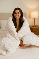 Eine jünge Frau sitzt auf dem Bett und kuschelt sich in unsere warme Bettdecke Schurwolle Lux von Wool-Line. Sie ächelt entspannt in die Kamera und fühlt sich in der Decke sichtlich wohl. 