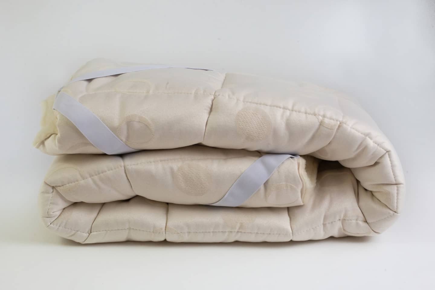 Das Schurwolle Unterbett Lux zusammengelegt auf weißem Untergrund. An der Seite sind zwei der weißen Spanngurte zu sehen, mit denen das Unterbett aus Schurwolle an die Matratze befestigt werden kann. 