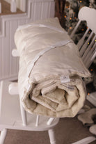 Der Bio-Topper aus Schafschurwolle Gold/Lux zusammengerollt auf einem weißen Schaukelstuhl. Im Hintergrund ist dezente Weihnachtsbeleuchtung zu sehen. 