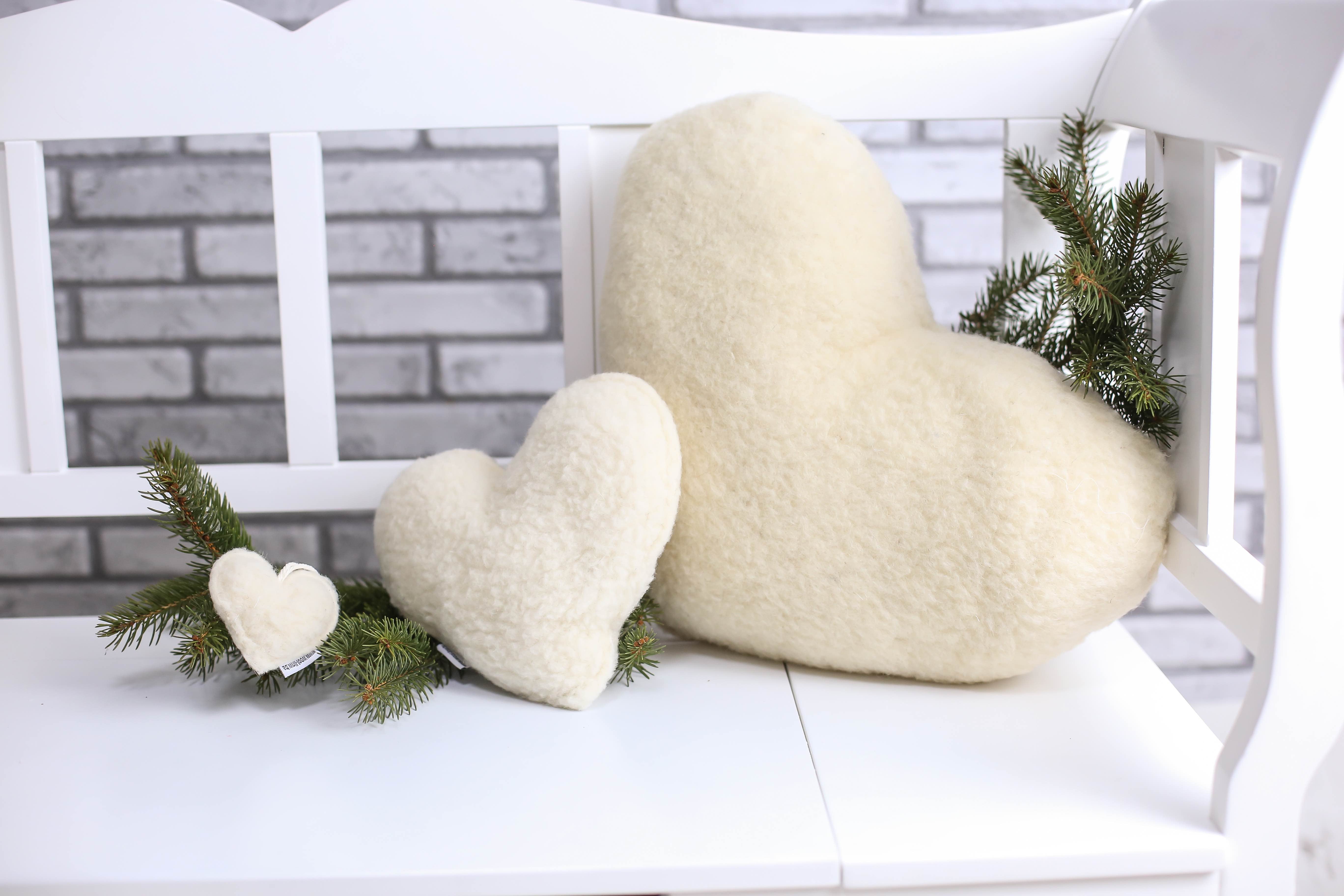 Drei Kissen in Herzform liegen nebeneinander auf einer weißen Outdoor-Bank. Links ein kleines Herzkissen, ein mittelgroßes in der Mitte und rechts ein großes Kissen in Herzform. Ein grüner Ast ist im Hintergrund als Dekoration abgelegt. 