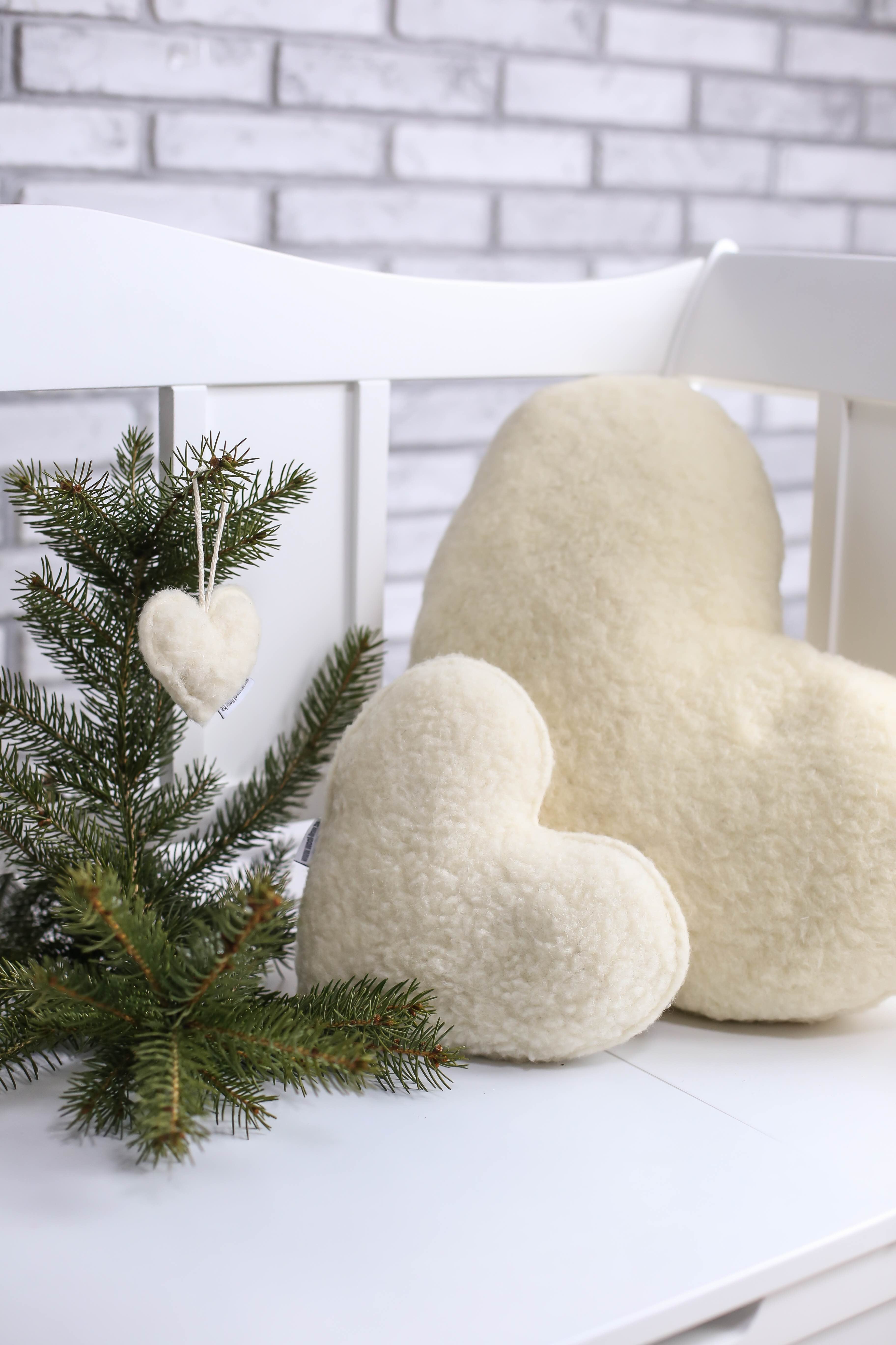 Das Kuschelherzkissen in drei verschiedenen Größen aufgereiht auf einer weißen Gartenbank. Das kleinste Kissen hängt an einem grünen Nadelbaumzweig. Das mittelgroße Herzkissen ist direkt vor dem größten Kuschelkissen Herz aufgestellt. 