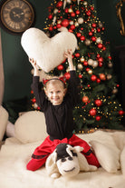 Ein Mädchen sitzt vor einem bunt geschmückten Weihnachtsbaum und hält glücklich das Herz-Kuschelkissen von Wool-Line in die Höhe. Es scheint so, als habe sie das Herzkissen zu Weihnachten geschenkt bekommen. 