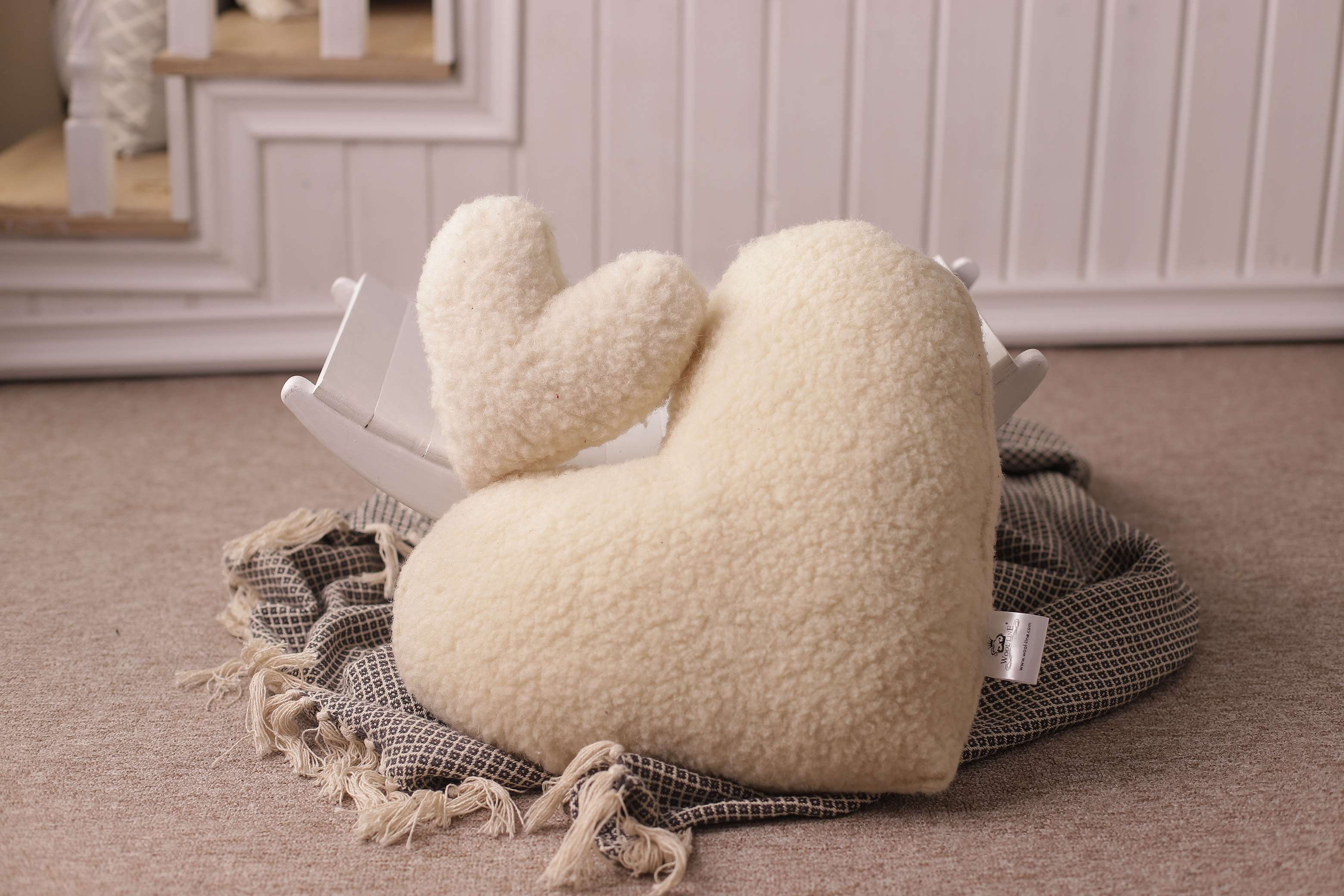 Zwei Kissen in Herzform von Wool-Line ineinander auf einer schwarz-weiß karierten Decke abgelegt. Das große Kissen nimmt einen großen Teil des Bildes ein, während ein kleineres Kissen in Herzform auf dem größeren Kuschelkissen abgestellt ist. 