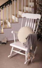 Das Kissen rund von Wool-Line auf einem weißen Schaukelstuhl gelegt. Es dient als weiche und angenehme Stütze für den Rücken. An der rechten Lehne ist eine Wolldecke aufgehängt. 