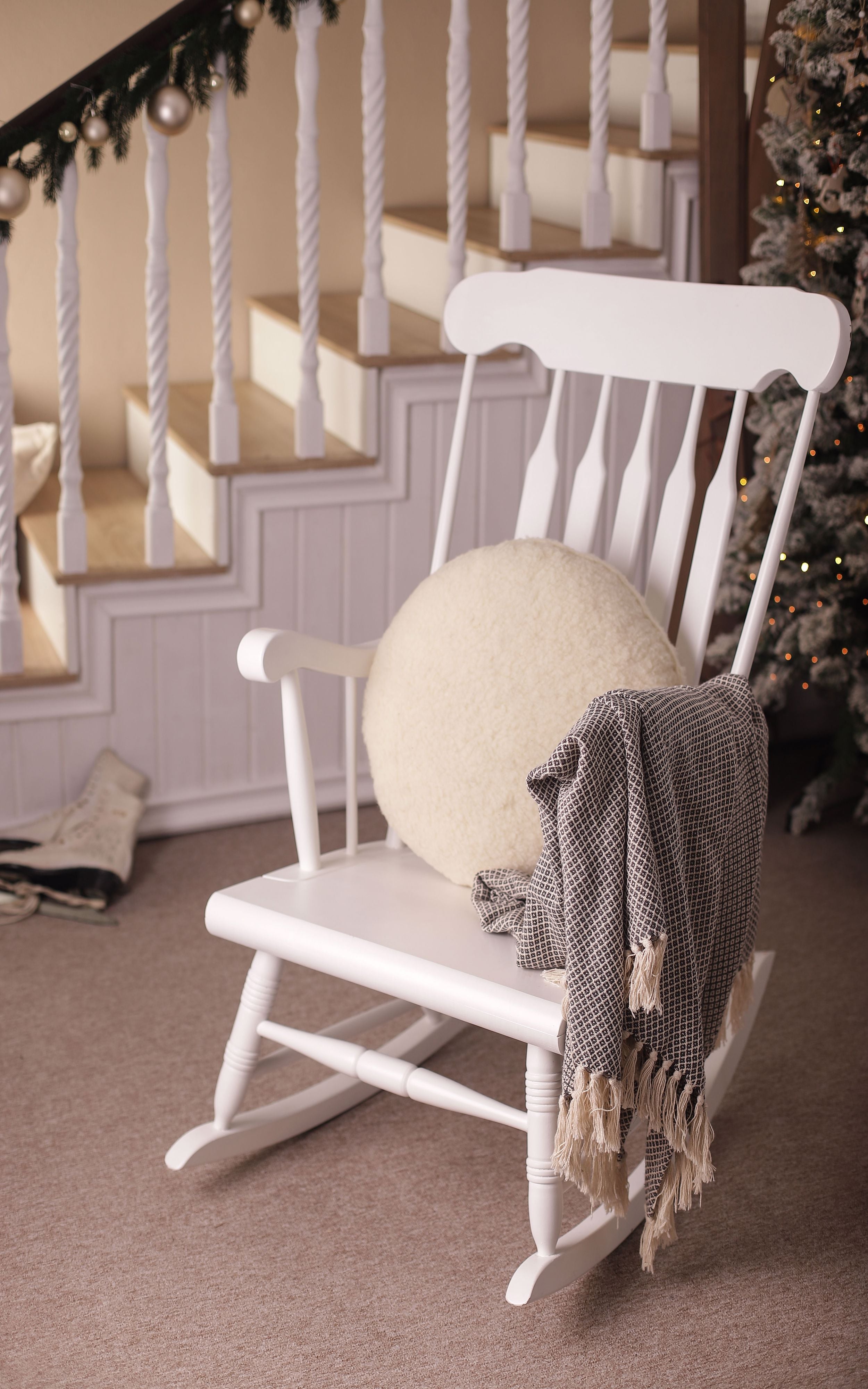 Das Kissen rund von Wool-Line auf einem weißen Schaukelstuhl gelegt. Es dient als weiche und angenehme Stütze für den Rücken. An der rechten Lehne ist eine Wolldecke aufgehängt. 