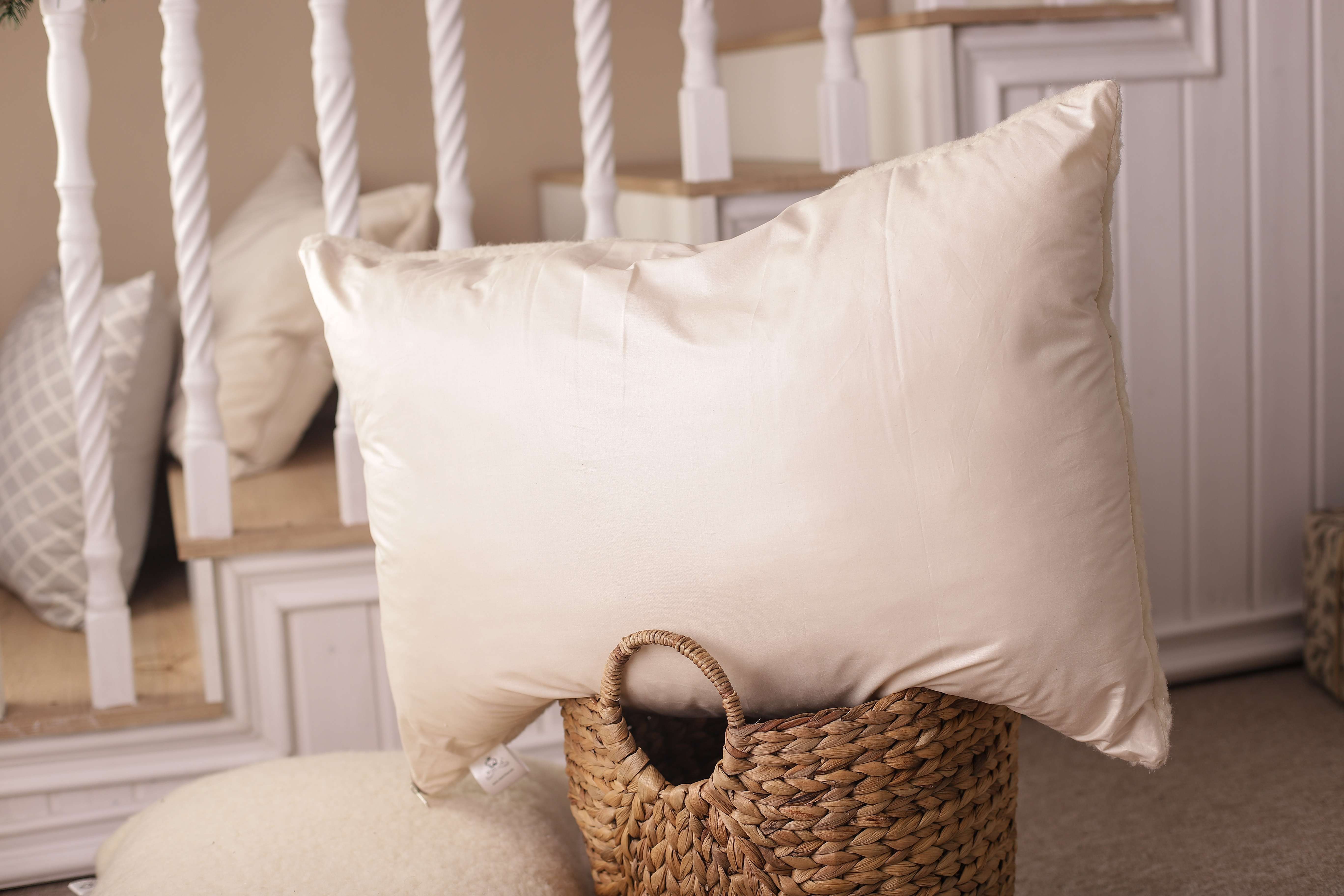 Das Kopfkissen Schafwolle Gold/Lux tront auf einem kleinen runden Korb. Zu sehen ist die glatte Seite aus Bio-Baumwolle, die besonders im Sommer für kühlende Entlastung sorgt. Im Hintergrund befindet sich eine Treppe, auf der zwei weitere Gold/Lux-Kissen aus Schafwolle liegen. 