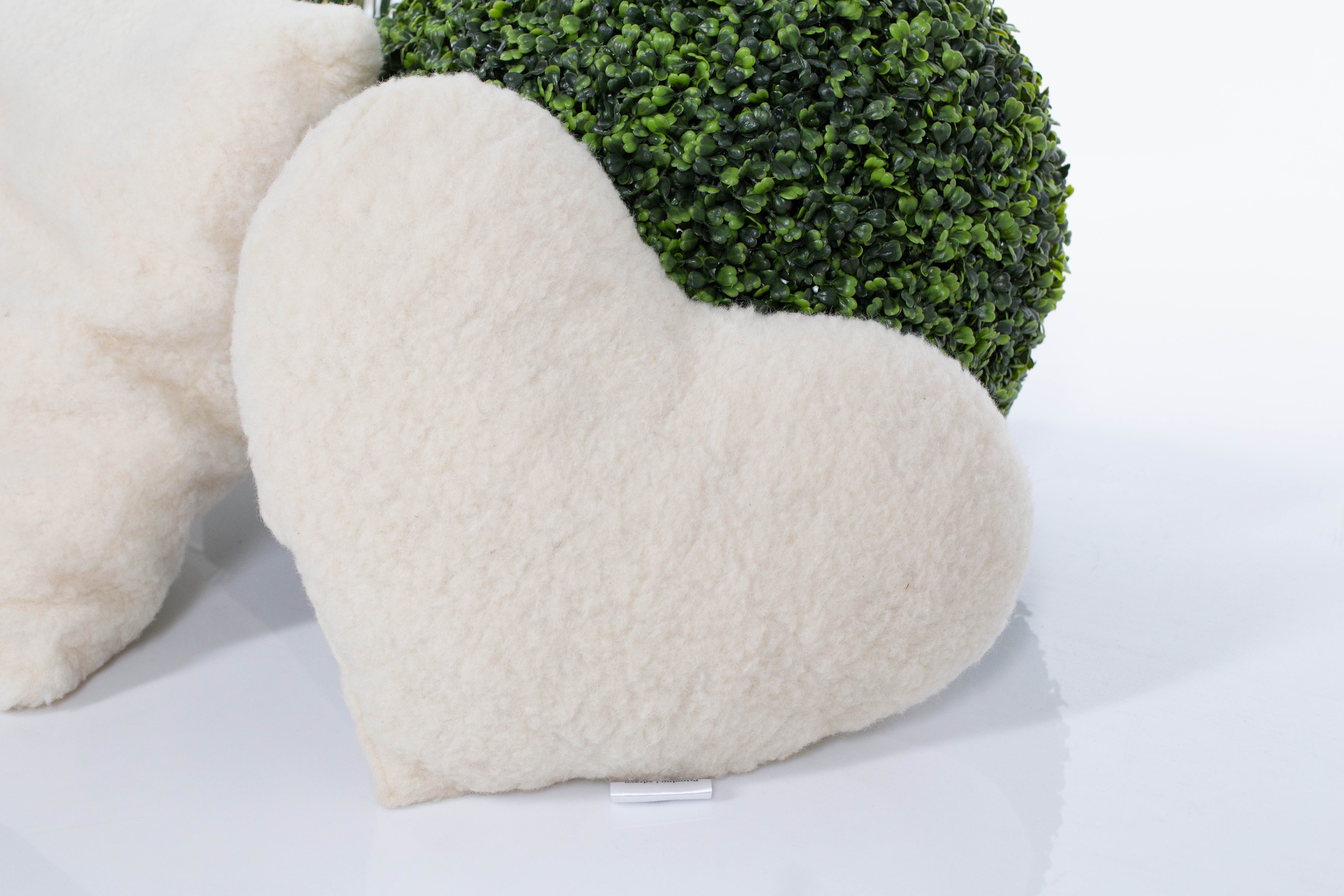 Das weiße Kuschelherz Kissen von Wool-Line auf weißem Untergrund fotografiert. Im Hintergrund befindet sich eine runde, güne Pflanze. Der weiche Filz des Herzkissens ist detailliert und gut ausgeleuchtet zu sehen. 