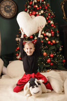 Ein etwa zehnjähriges Mädchen reckt das Kuschelkissen Herz von Wool-Line freudestrahlend vor einem bunt geschmückten Weihnachtsbaum in die Höhe. 