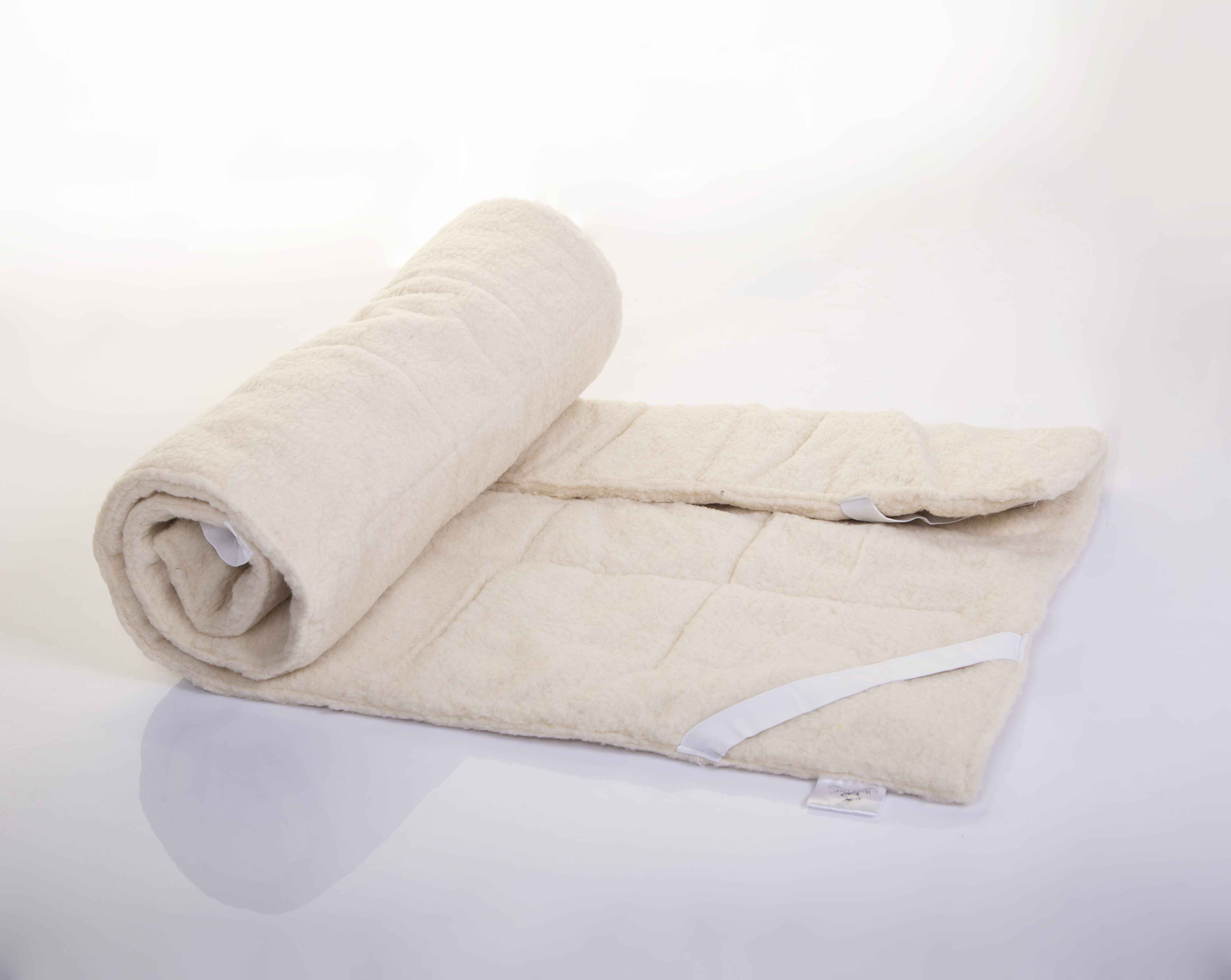 Die Matratzenauflage Schurwolle Gold von Wool-Line auf weißem Untergrund. Die linke Seite ist flexibel eingerollt, die andere Seite liegt teilweise zusammengelegt flach auf dem Untergrund. Dank der Schafschurwolle im Futter der Matratzenauflage ist das Produkt sehr elastisch ohne sichtbare Dellenbildung. 