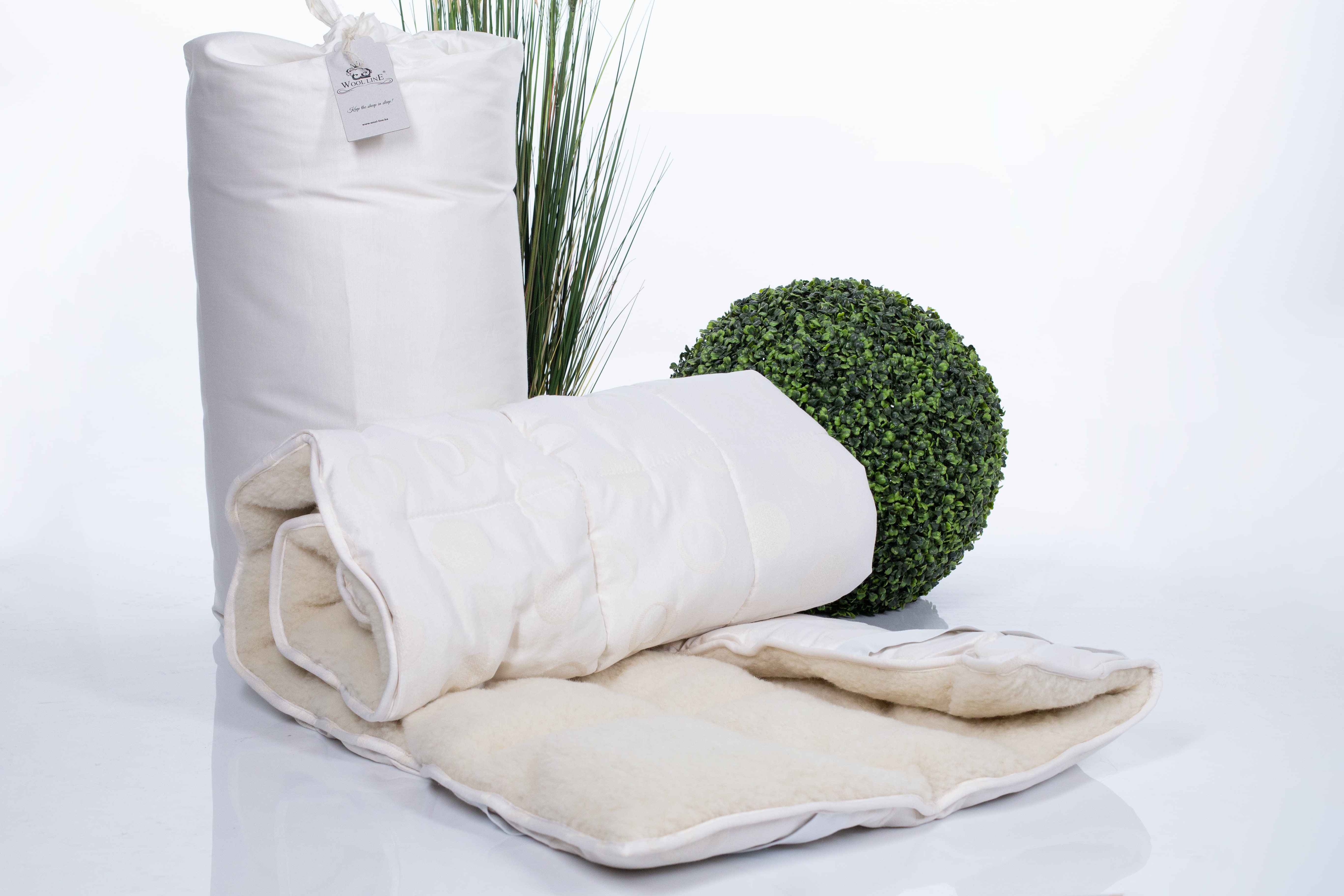 Der Matratzentopper Natur Gold/Lux eingerollt auf weißem Untergrund. Im Hintergrund befinden sich zwei Pflanzen und ein Wool-Line Paket ohne Plastik fertig zum Versand. 