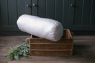 Die Nackenrolle Bio von Wool-Line auf einer kleinen dunkelbraunen Holzkiste ausgestellt. Die Nackenrolle ist mit dem Bezug aus Bio-Baumwolle bezogen und mit echter Schafschurwolle gefüllt. 