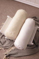 Die Nackenrolle beige Gold und die Nackenrolle weiß mit Bio-Baumwollbezug von Wool-Line liegen nebeneinander auf einer kleinen Ablage. Darunter befindet sich eine schwarz-weiß karierte Decke. 