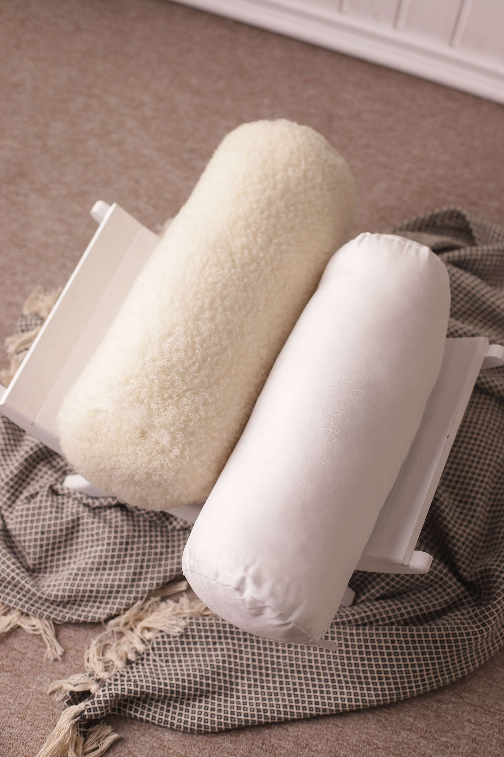 Die Nackenrolle beige Gold und die Nackenrolle weiß mit Bio-Baumwollbezug von Wool-Line liegen nebeneinander auf einer kleinen Ablage. Darunter befindet sich eine schwarz-weiß karierte Decke. 