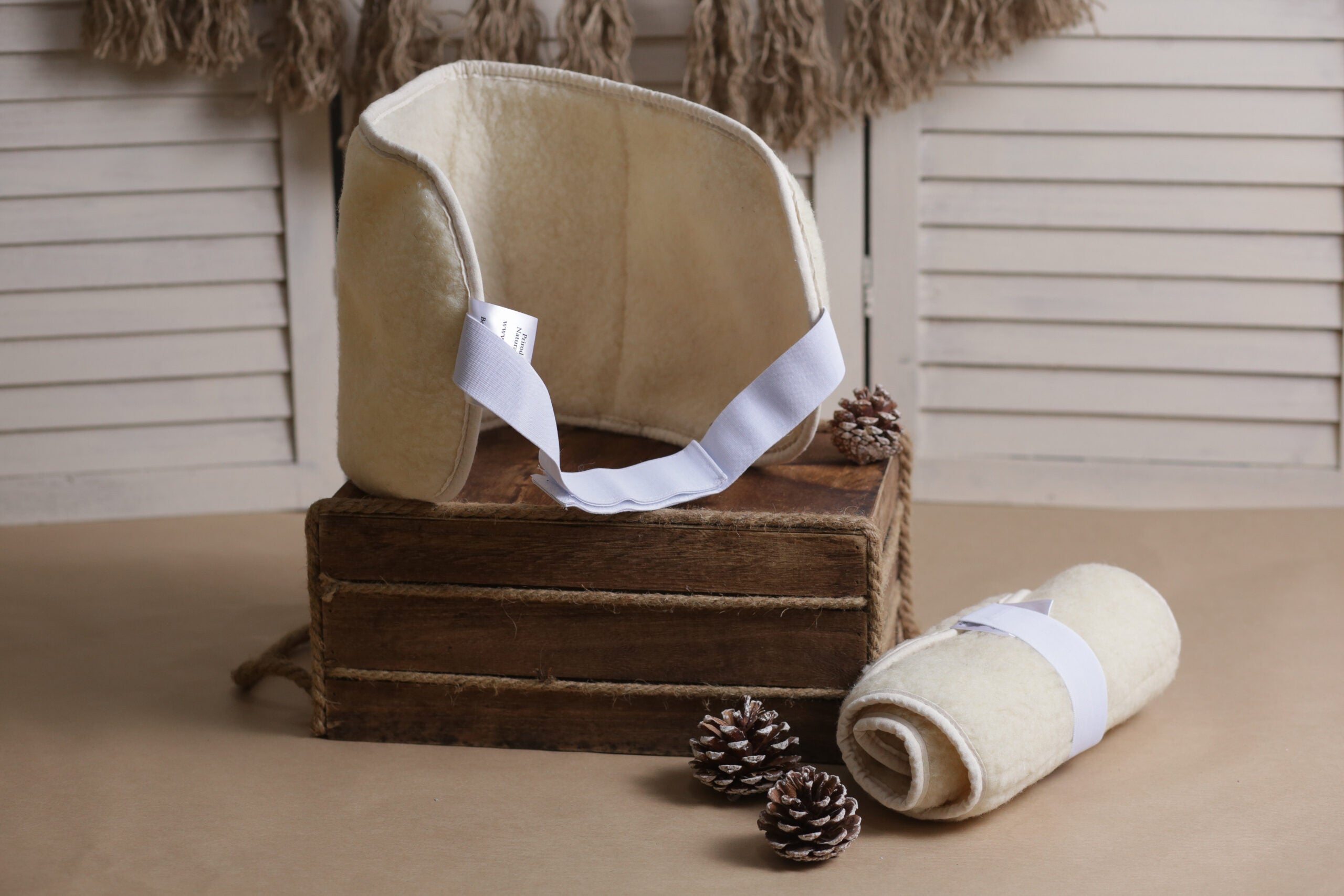 Die Rückenstütze als Gürtel von Wool-Line auf einer dunkelbraunen Anhöhe aus Holz. Um den Rückengürtel herum befinden sich drei Tannenzapfen und eine zweite Rückenstütze als Gürtel, die komplett eingerollt und mit dem Klettverschluss fixiert ist. 