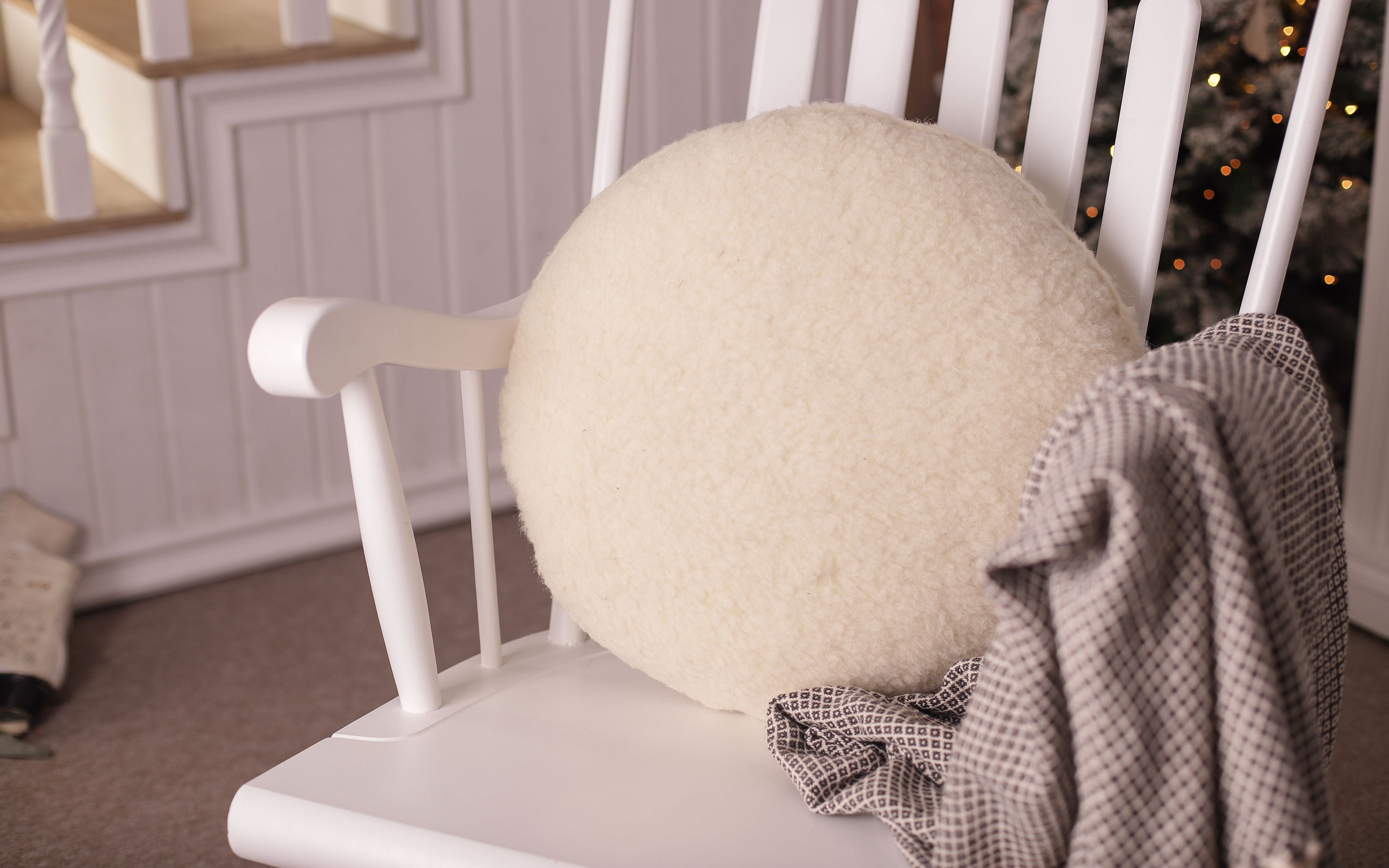 Das Rundkissen von Wool-Line auf einem weißen Schaukelstuhl. Das runde Kissen ist an die Lehne angelehnt und dient als weiche Rückenstütze. 