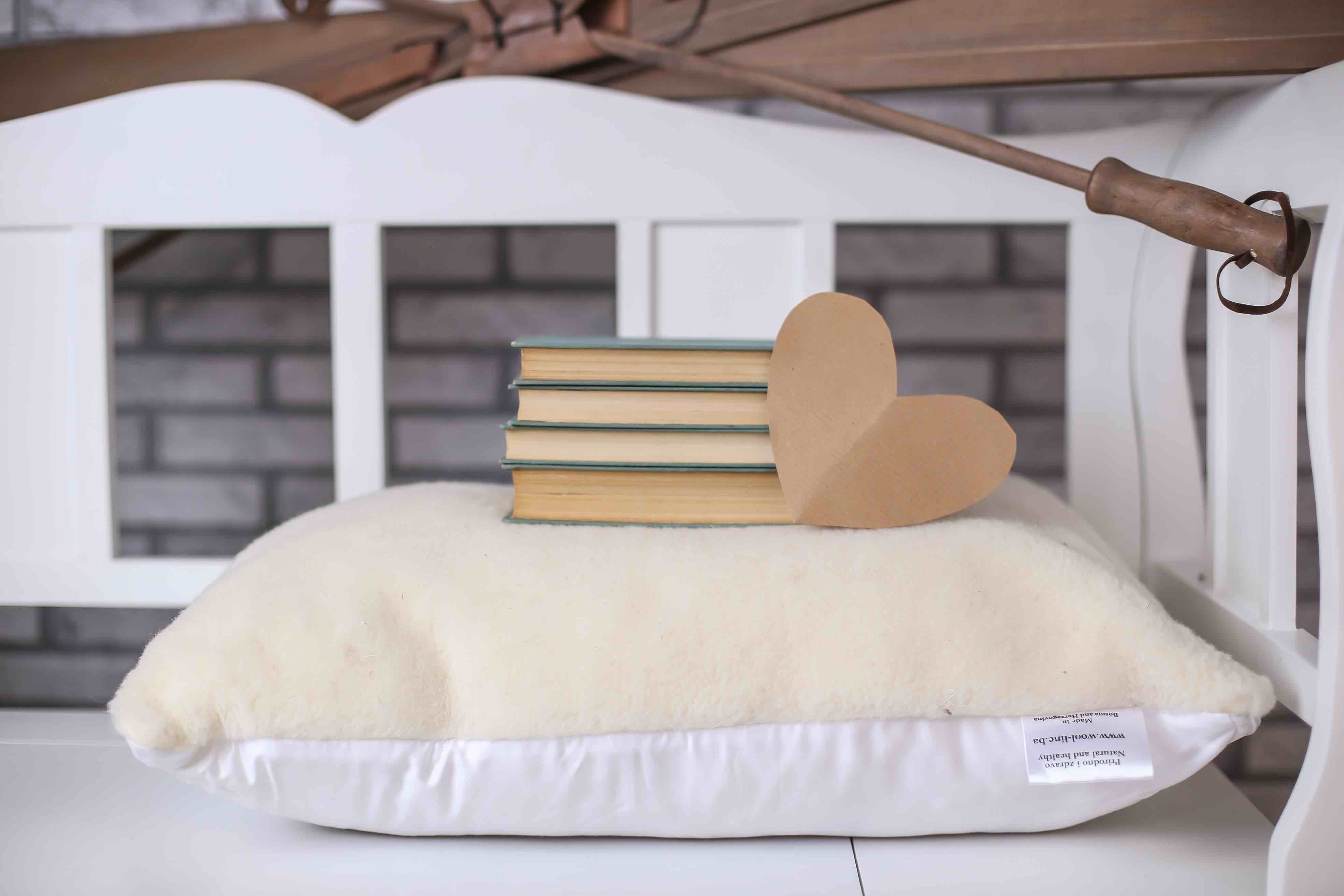 Das Schafwolle Kissen Gold/Lux von Wool Line befindet sich auf einer weißen Ablage. Auf der Oberseite aus weichem Filzbezug sind vier Bücher gestapelt. Davor liegt ein kleines, braunes Papierherz. 