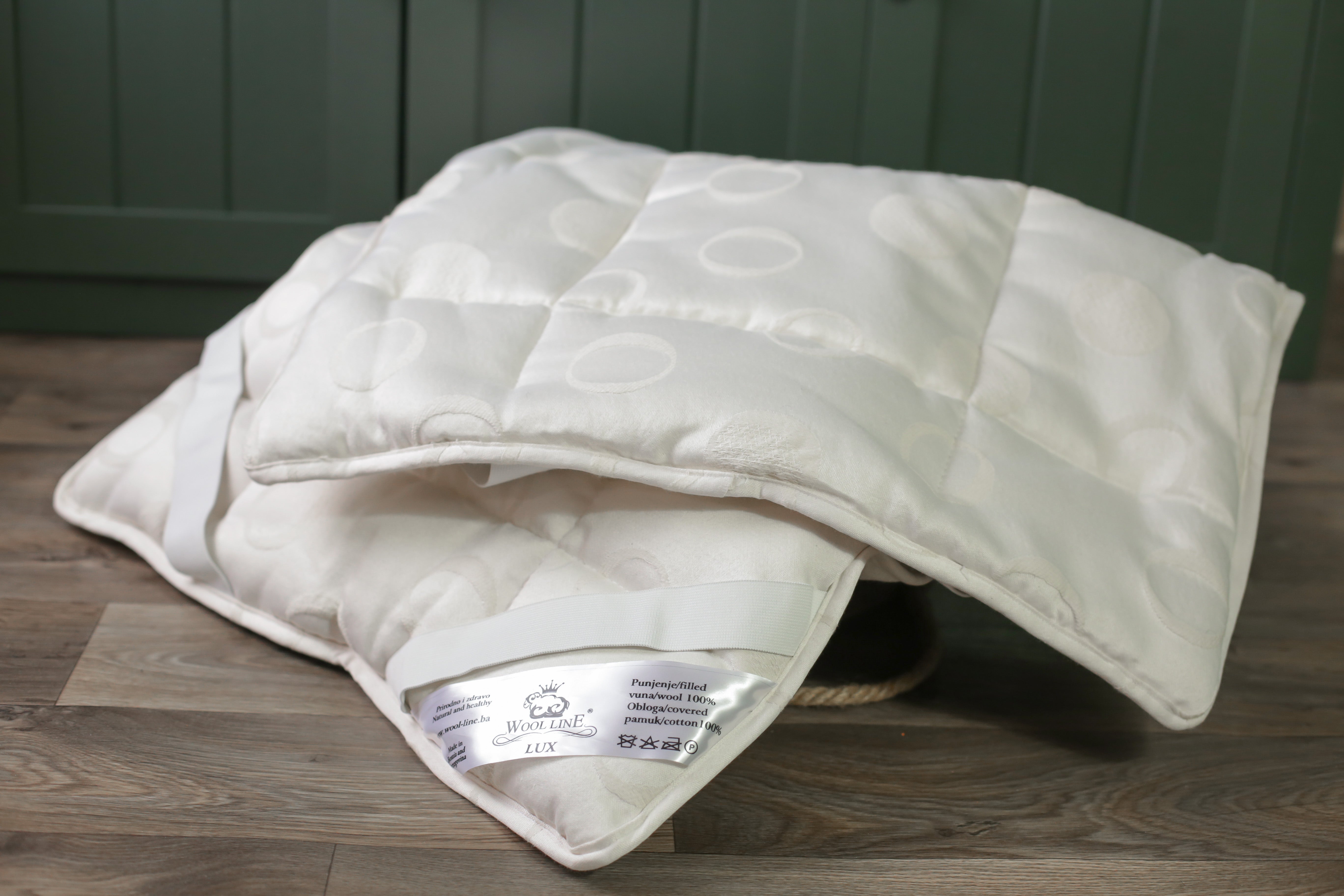 Das Schurwolle Unterbett Lux von Wool-Line auf einer dunkelbraunen Ablage aus Holz. Das Unterbett ist eingeklappt. Am unteren Eck kommt ein Zettel zum Vorschein, auf dem die Pflegehinweise des Schurwoll-Unterbetts und das Wool-Line Logo zu sehen sind. 