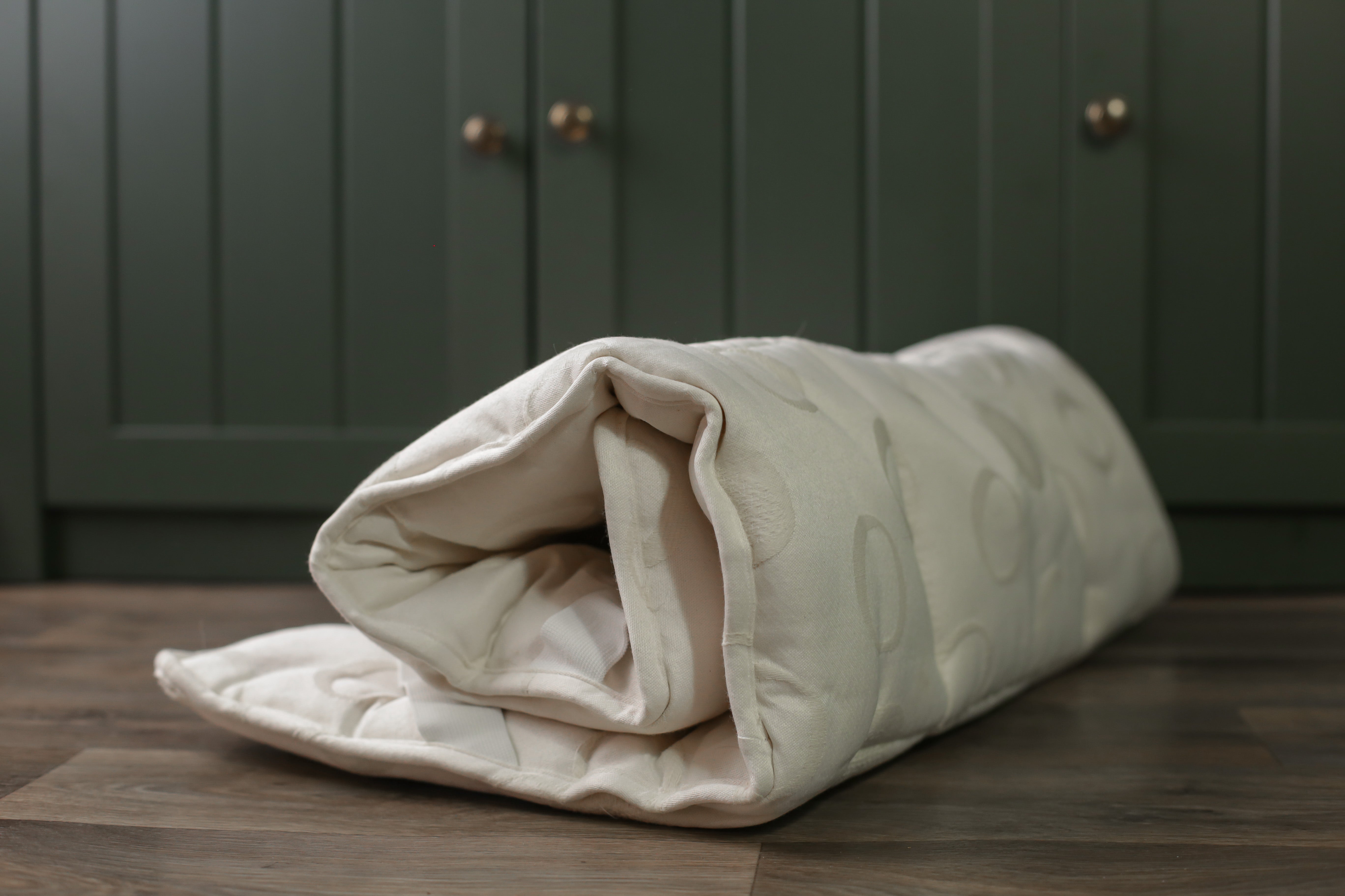 Unser Wool-Line Unterbett aus Schurwolle auf einer Ablage aus dunkelbraunem Holz. Das Unterbett ist eingerollt und zeigt die gesamte Biegsamkeit ohne Dellenbildung. 