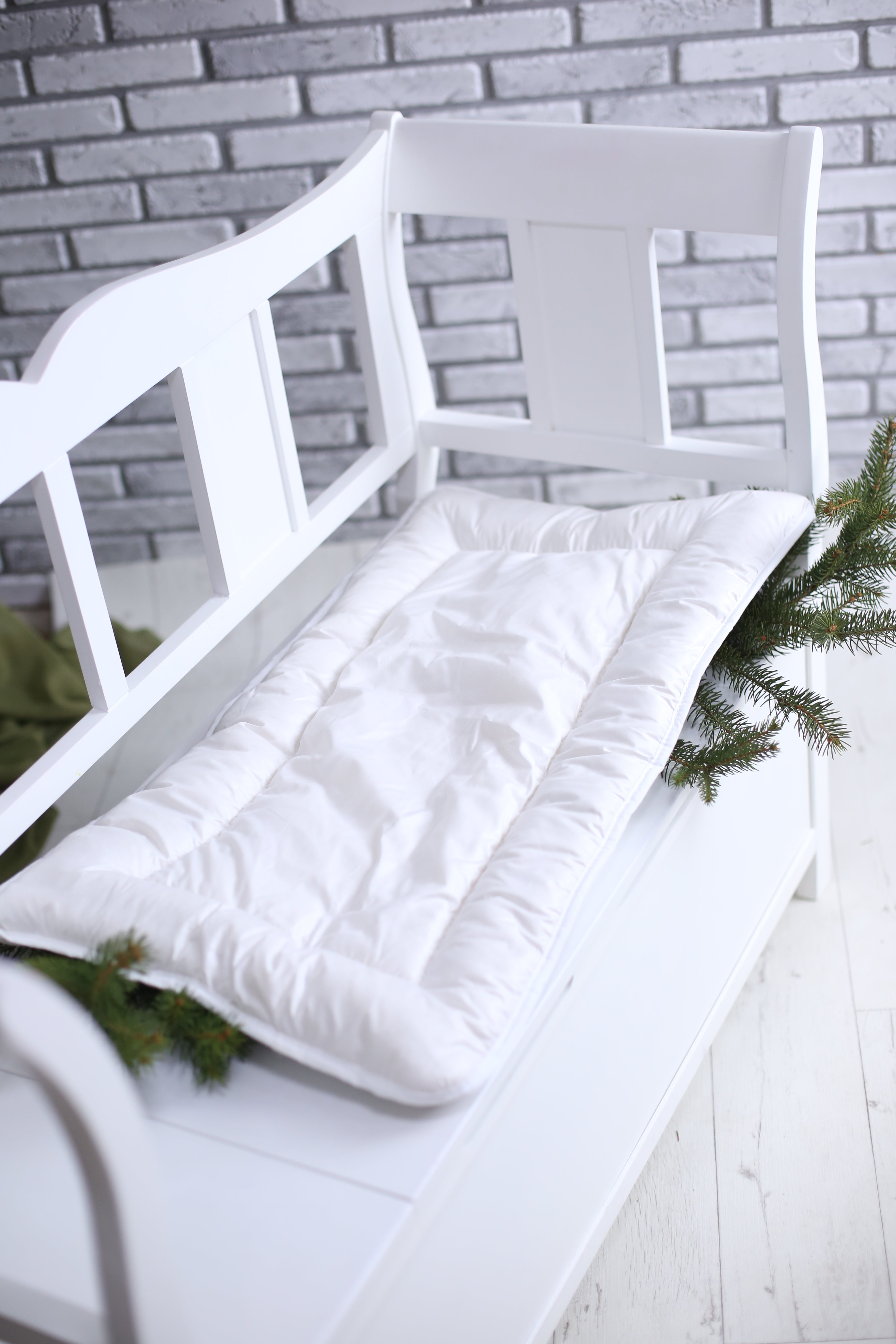 Die Wickelauflage für Babybett von Wool-Line mit echter Schafschurwolle gefüllt auf einer weißen Bank. Darunter liegen grüne Äste, was die Natürlichkeit der Wickelauflage und die vielseitige Verwendung verdeutlichen soll. 